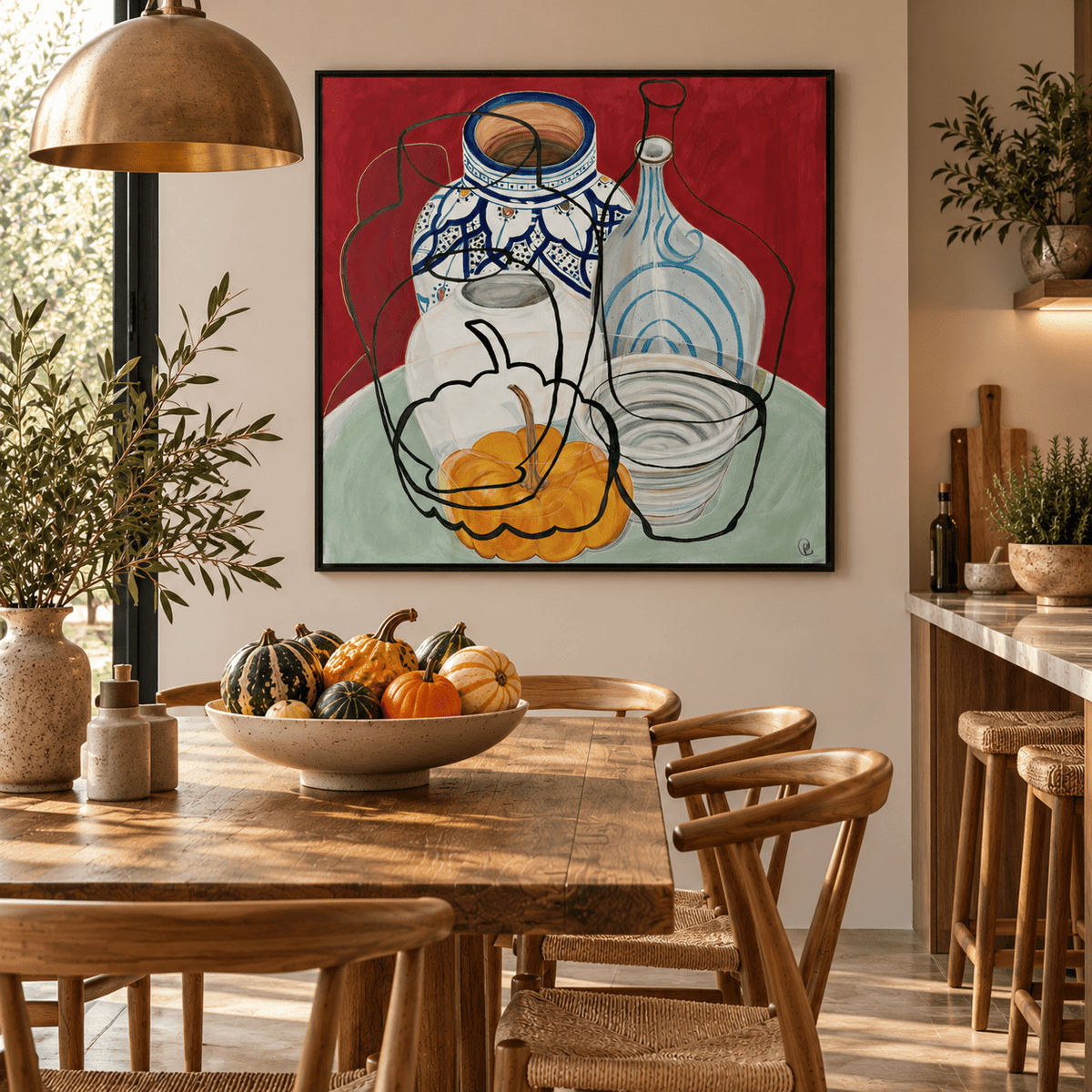 A framed abstract still life painting by Rebecca Carr hangs on a beige wall above a wooden dining table in a styled kitchen-dining room. The artwork features a red background with blue, white, black, and yellow-orange painted glass bottles and a bowl.