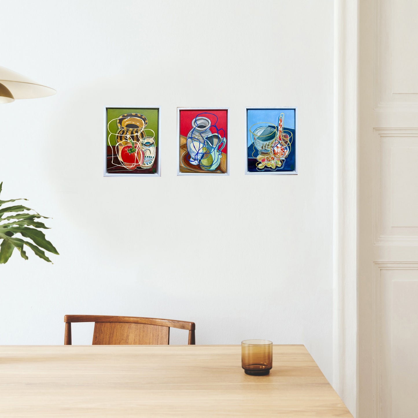 Three colourful still life paintings by artist rebecca carr on a white wall above a wooden table.
