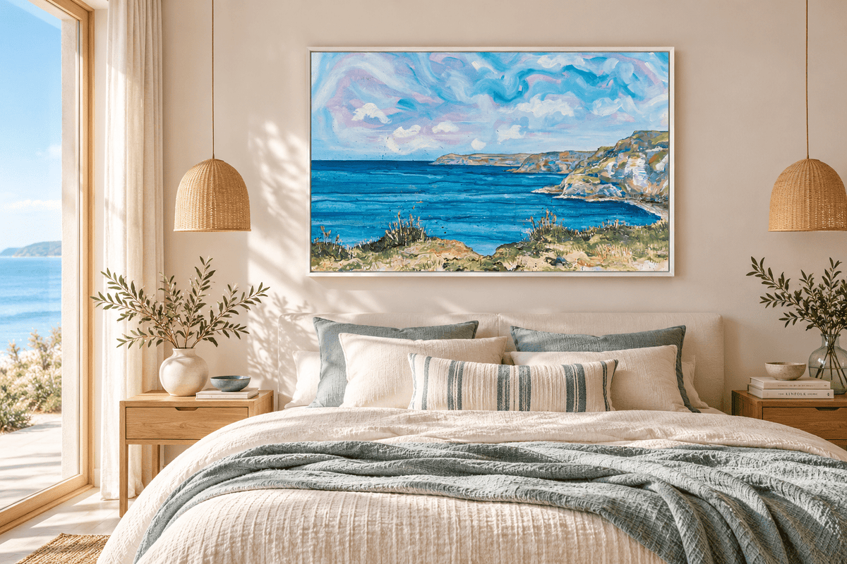 A framed landscape painting of a coastal bay with blue water, green cliffs, and a cloudy sky, displayed above a neatly made bed in a bright, modern bedroom.