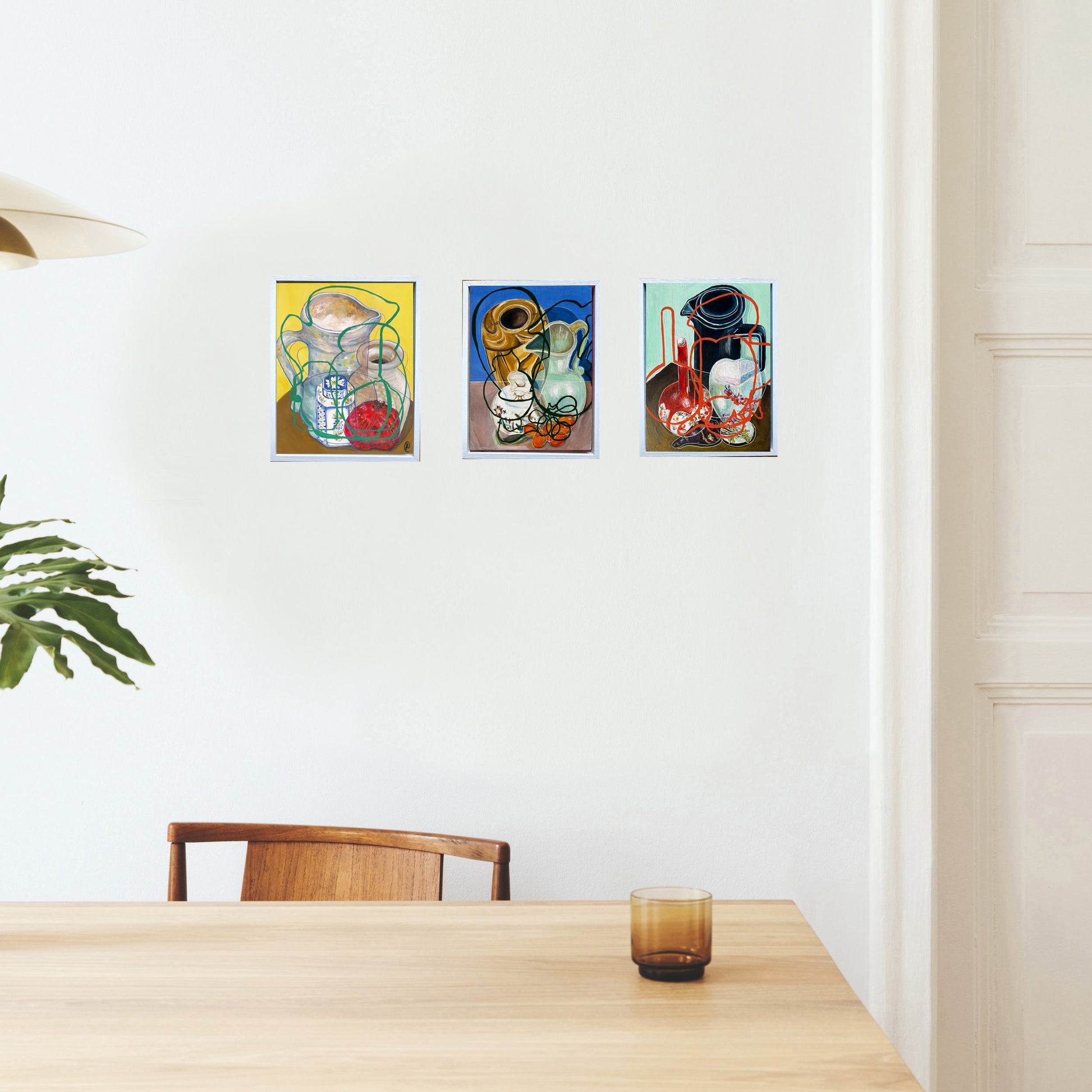 Three colorful paintings on a white wall above a wooden table. Still life by Rebecca Carr