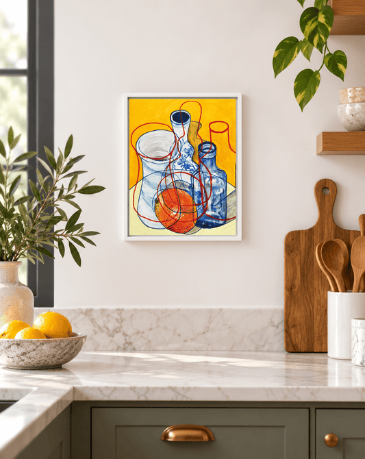 A framed art print featuring a colorful still life of glass bottles on a yellow background, hanging on a white kitchen wall above a marble countertop with lemons and wooden cutting boards.