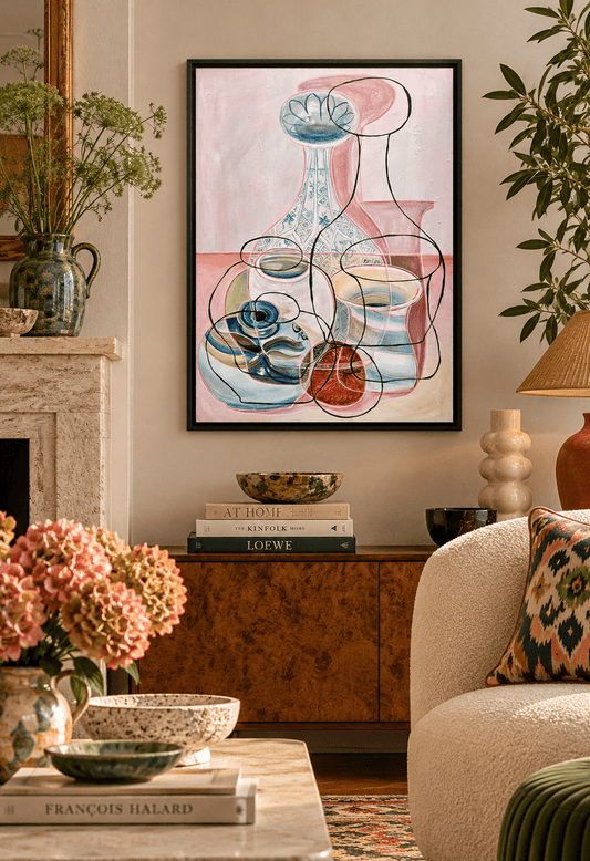 A framed pink abstract still life painting hangs on a beige wall in a styled living room. The artwork features a pink and white background with blue, green, and red geometric shapes and line art.