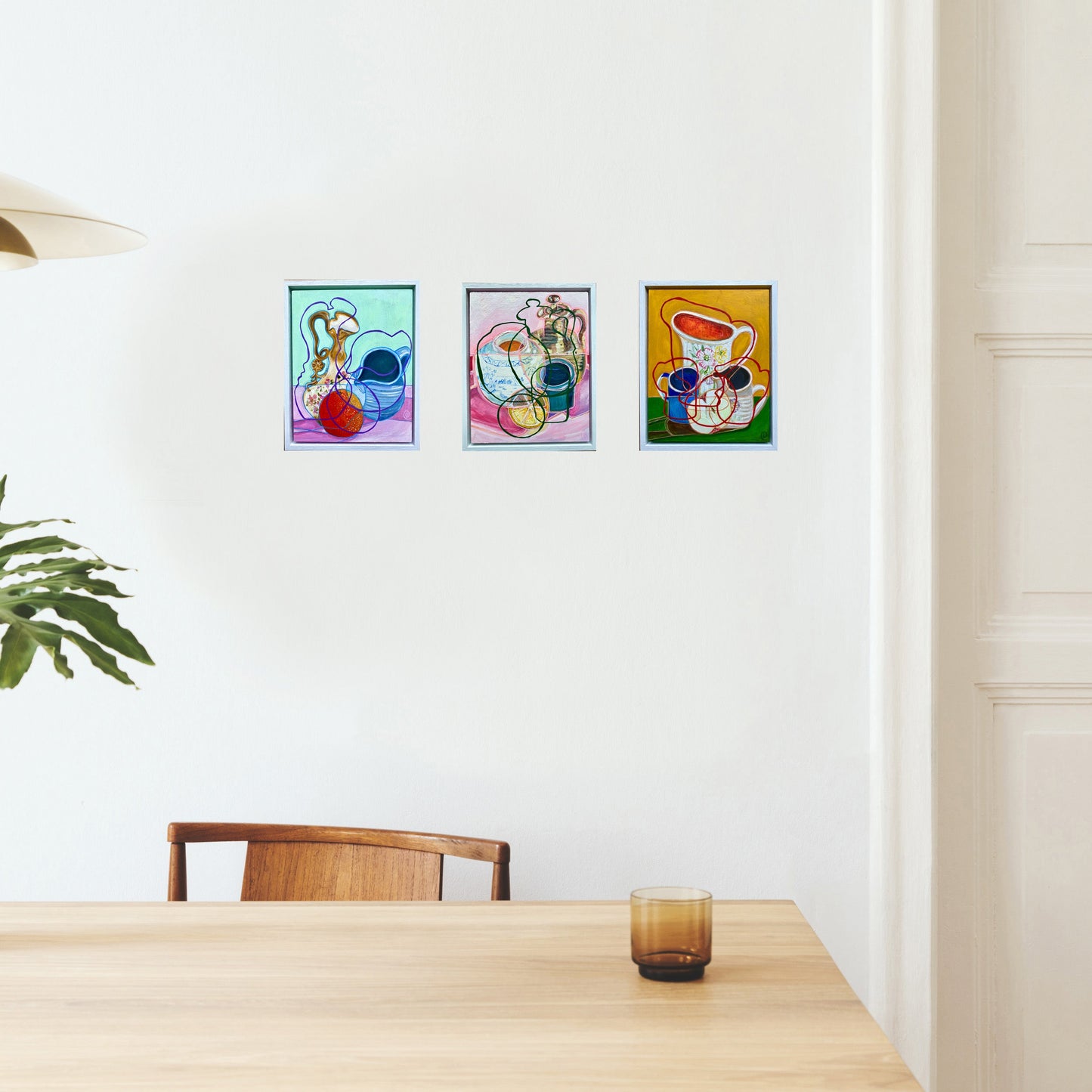 Three colourful still life paintings by rebecca carr artist on a white wall above a wooden table.