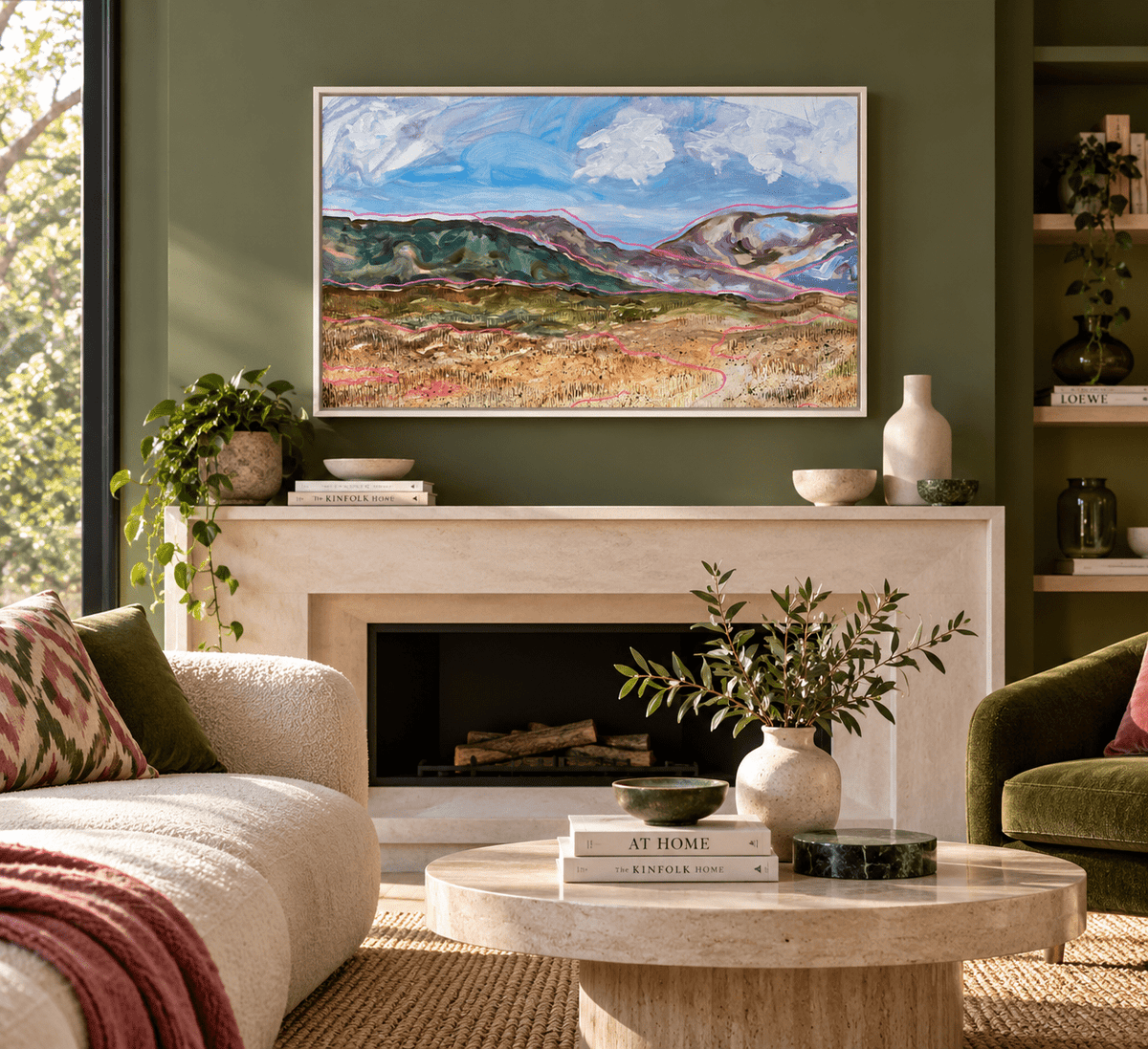 A framed landscape painting of mountains and a field hangs above a fireplace in a styled living room with a sofa, coffee table, plants, and shelving.