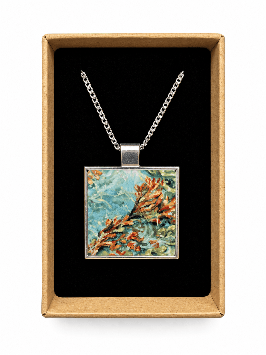 A square pendant necklace displayed inside an open kraft gift box. The pendant features a colorful floral or botanical painting with blue, green, orange, and beige tones, suspended from a silver-toned chain against a black interior background.